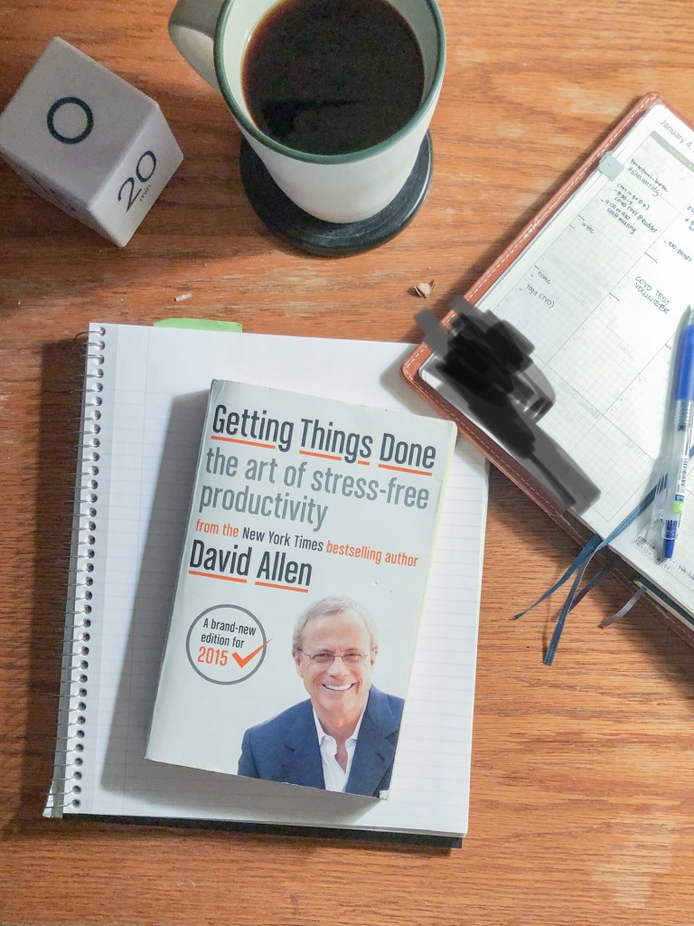 A desktop with a note book, a planner, a cup of coffee and a copy of David Allen's book Getting Things Done